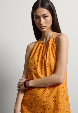 Jonge vrouw met lang donker haar die een oranje mouwloze jurk draagt met bloemmotieven en een rode armband, staande tegen een effen achtergrond.