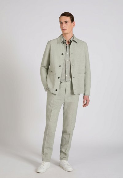 Mężczyzna stojący, ubrany w jasnoszarą zapinaną na guziki kurtkę, dopasowane spodnie oraz białe trampki, patrzący nieco w bok na jednolitym tle.