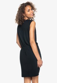 Vestido negro sin mangas con cintura fruncida, textura suave, hasta la rodilla y escote redondo, visto desde la espalda con cabello ondulado.