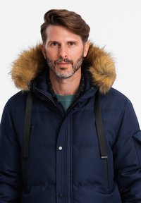 Giacca puffer blu navy con cappuccio foderato in pelliccia, dotata di tasca con zip, cinturini regolabili e una texture morbida. Visibili dettagli di cucitura robusta.