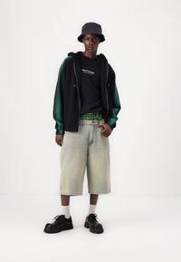Svart hettegenser med grønne detaljer, svart t-skjorte med hvit tekst, lyse denimshorts, svarte sko, grå sokker og svart bøttehatt.