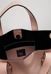 Růžová taška přes rameno s černým interiérem, hladkým povrchem, zlatými kovovými akcenty a viditelnou značkovou etiketou uvnitř. Design bez zapínání.