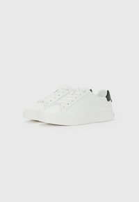 Sneaker bianche basse con lacci bianchi e linguetta nera sul tallone, esposte su uno sfondo bianco.