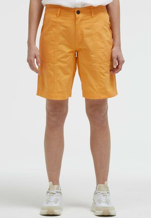 Person trägt knallorange knielange Shorts mit aufgesetzten Taschen, weiße Sneakers und ein in die Hose gestecktes weißes Hemd, stehend vor einfarbigem Hintergrund.