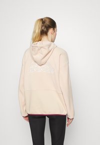 Světle růžová mikina s kapucí a volným střihem, měkká na dotek, s bílým logem adidas na zadní straně. V kombinaci s tmavými legínami.