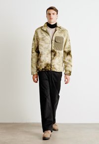 Fleece jack in beige en groene camo-print, met een borstzak, ritssluiting en zachte textuur, gecombineerd met zwarte broek en schoenen.