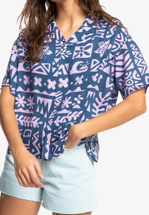 Frau trägt ein kurzärmeliges Hemd mit blau-pinkem Muster und hellblaue Jeans-Shorts, eine Hand in der Tasche, vor weißem Hintergrund.