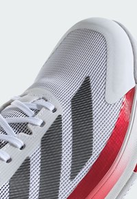 Baskets blanches avec une tige en mesh texturé, accents gris et une bande latérale rouge. Équipées de lacets plats et d'un design de bout arrondi.