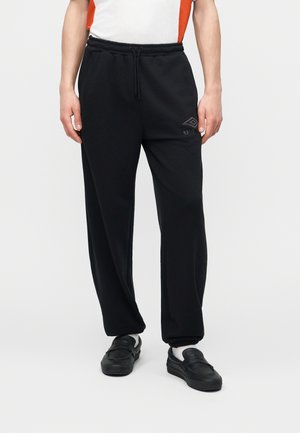 Άτομο που φοράει μαύρο παντελόνι φόρμας Umbro με κορδόνι στη μέση, λευκές κάλτσες, μαύρα παπούτσια χωρίς κορδόνια και λευκό και πορτοκαλί τοπ, στέκεται σε λευκό φόντο.