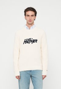 Κρεμ χρώματος πλεκτό πουλόβερ με ριγωτή υφή και ναυτικό λογότυπο που γράφει "Hilfiger" έντονα εκτυπωμένο στο στήθος.