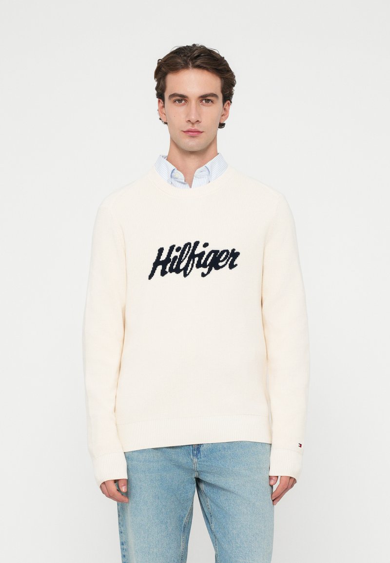 Krém színű kötött pulóver, bordázott textúrával, a mellkasán feltűnően elhelyezett "Hilfiger" felirattal, sötétkék színben.