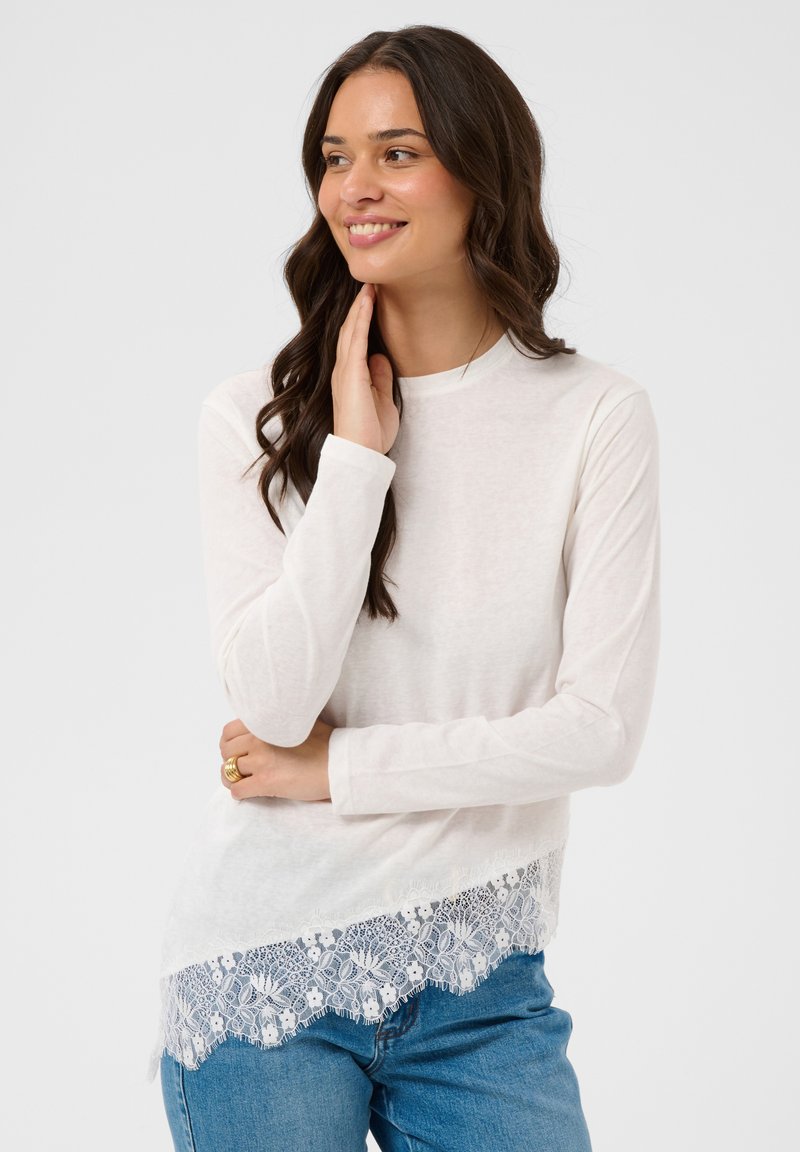 Femme aux longs cheveux bruns portant un haut blanc à manches longues avec une bordure en dentelle et un jean bleu, souriante et se touchant le visage, debout devant un fond uni.