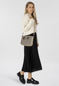 Beige geometrisch patroon handtas met dubbele handvatten en een verstelbare schouderband, afgezet tegen een model in een crèmekleurige trui en een zwarte plissékilt.