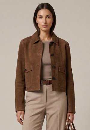 Frau trägt eine braune Wildlederjacke mit Knöpfen über einem beigen, gerippten Top, beige Hose mit braunem Gürtel, hält eine braune Handtasche.