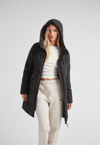 Kvinde iført en sort hættejakke med quiltet mønster over en hvid crop top og beige sweatshirtbukser foran en ensfarvet lys baggrund.