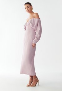 Dlouhé, ne ramenní levandulové šaty s volánkovým korpusem a přiléhavým pasem, s dlouhými rukávy a hladkou saténovou texturou. V páru se zlatými páskovými podpatky.