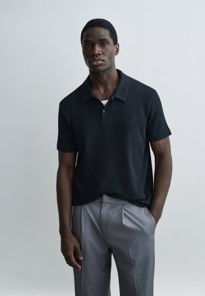 Hombre joven de piel oscura vistiendo una camiseta tipo polo negra de manga corta y pantalones grises con pliegues, de pie frente a un fondo liso gris claro.