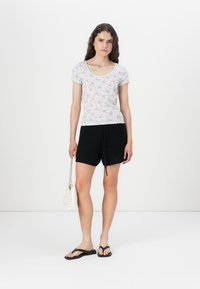 Top a maniche corte con motivo floreale, di colore chiaro; pantaloni corti neri con texture e coulisse; borsa bianca; infradito nere.