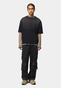 Camiseta negra holgada superpuesta sobre una camiseta de manga larga beige, combinada con pantalones cargo oscuros con múltiples bolsillos y detalles texturizados.