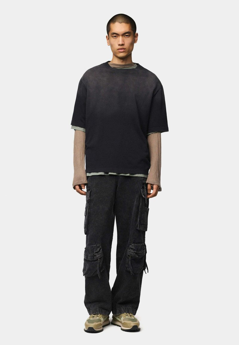 Camiseta negra holgada superpuesta sobre una camiseta de manga larga beige, combinada con pantalones cargo oscuros con múltiples bolsillos y detalles texturizados.