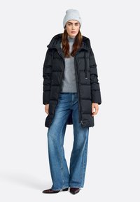 Zwart gewatteerd jasje met capuchon, voorrits en verstelbare koorden. Draagt over een grijze trui en wijde jeans. Grijze gebreide beanie.