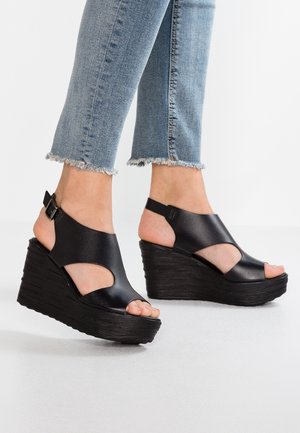 Jalat sinisissä lyhyissä farkuissa, joissa on mustat nahkaiset kiilakantaiset sandaalit, joissa on avoimet varpaat ja nilkkahihnat valkoisella lattialla.