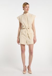 Rochie bej fără mâneci, cu guler înalt, talie croitică și pantaloni scurți. Fabricată dintr-un material texturat. Modelul poartă tocuri nude.