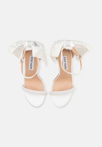 Sandales blanches à talons hauts avec une sangle en perles, un nœud en tissu scintillant et des accents argentés. Texture lisse avec une semelle intérieure nude et une fermeture à boucle.