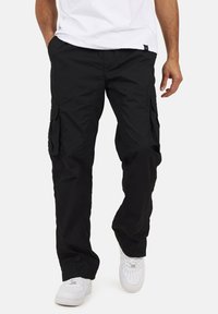 Czarne spodnie cargo o luźnym kroju, z wieloma bocznymi kieszeniami i elastycznymi mankietami. Noszone z białą koszulką i białymi trampkami.