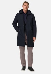 Temno moder plašč srednje dolžine s kapuco, ki ima zadrgo in stranske žepe, nosi se preko srajce z gumbi in svetlih hlač.