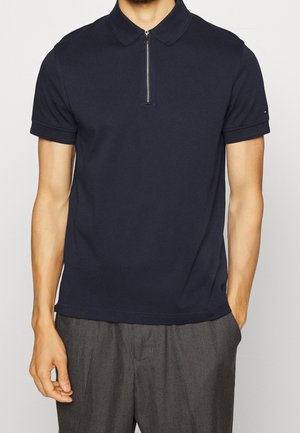 Man draagt een marineblauw polo met korte mouwen en rits aan de voorkant en grijze broek, handen ontspannen langs zijden tegen een effen achtergrond.
