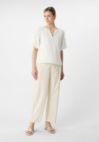 Weiße bestickte Bluse mit V-Ausschnitt und kurzen, ausgestellten Ärmeln, kombiniert mit leicht beigen, locker sitzenden Hosen und Riemchensandalen.