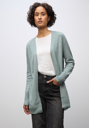 Lysblå cardigan lavet af blødt, strikket stof, med åben front, ribbede manchetter og en afslappet pasform, båret over en hvid skjorte og sorte bukser.