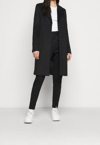 Dlouhý černý kabát s klopovým límcem, jednorušný střih, doplněný černými zúženými kalhotami a bílými teniskami. Celkově hladká textura.