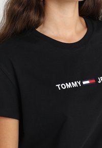 Camiseta de algodón negra con cuello redondo y mangas cortas, con el texto en blanco "TOMMY" y un acento de logo en rojo y azul.