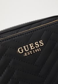 Sorte quilted syntetisk taske med guld "GUESS EST 1981" logo, der har vandret syninger og en glat tekstur.