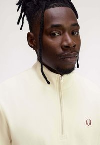 Felpa bianca sporco con zip e collo alto, con un piccolo logo rosso scuro sul petto e tessuto testurizzato.