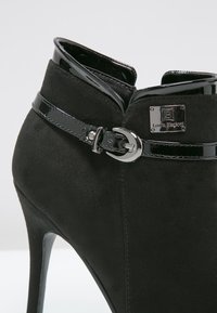 Czarne zamszowe botki za kostkę z akcentami z lakierowanej skóry i detalem w postaci klamry. Posiadają wysoki obcas w stylu stiletto oraz gładką teksturę.