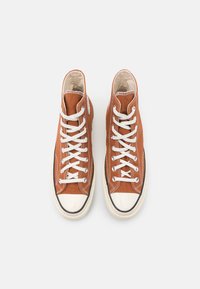 Bruna högsneakers med vita snören, tillverkade av canvasmaterial, med texturerad yta, gummikåpa vid tån och svarta detaljer längs sulan.