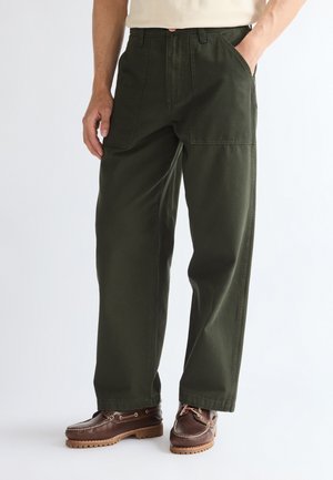 Homme portant un pantalon large vert foncé avec des poches plaquées à l'avant et des chaussures en cuir marron, debout avec une main dans la poche.