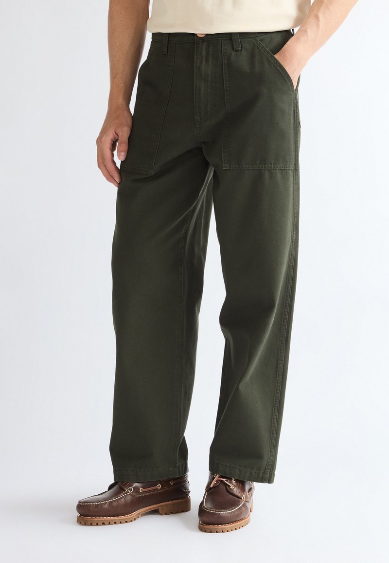 Homme portant un pantalon large vert foncé avec des poches plaquées à l'avant et des chaussures en cuir marron, debout avec une main dans la poche.