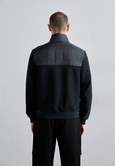 Black sweatshirt with a padded yoke, featuring a high collar, ribbed cuffs, and hem. Smooth fabric with a matte finish, minimalistic design.