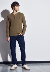 Olivgrön långärmad henley-tröja med texturerad tyg, knappdetalj vid halsringningen, kombinerad med mörkblå slim-fit jeans och krämfärgade sneakers.