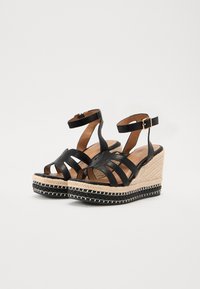 Sandales à talons en cuir noir avec plusieurs bandes, accents en paille naturelle et un talon texturé. Fermeture à boucle sur la sangle de cheville.