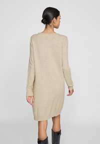 Beige gestricktes Sweatkleid mit langen Ärmeln, gerippten Bündchen und einem etwas längeren Rücken. Der Stoff wirkt weich und strukturiert.