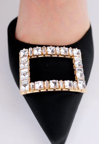 Scarpa nera a punta con una fibbia rettangolare dorata decorata con strass trasparenti, su uno sfondo di tessuto liscio.