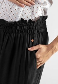 Schwarze, leichte Hose mit einem gerafften Taillengummi, die über einen plissierten Stoff verfügt und ein kleines Metallelement in der Nähe der Tasche hat.