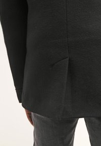 Svart ullblandad jacka med texturerad yta, som har en slits bak och en skräddarsydd passform, ihop med mörkgrå byxor.