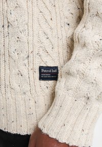 Kabelstickad tröja i krämfärg med spräcklig textur. Har ribbade muddar och nederkant. Taggen läser "Petrol Ind Denim Driven" i svart.