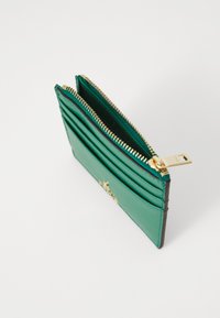 Grønn lærwallet med gullglidelås, flere kortlommer og en glatt tekstur. Har et dekorativt metallaccent på forsiden.